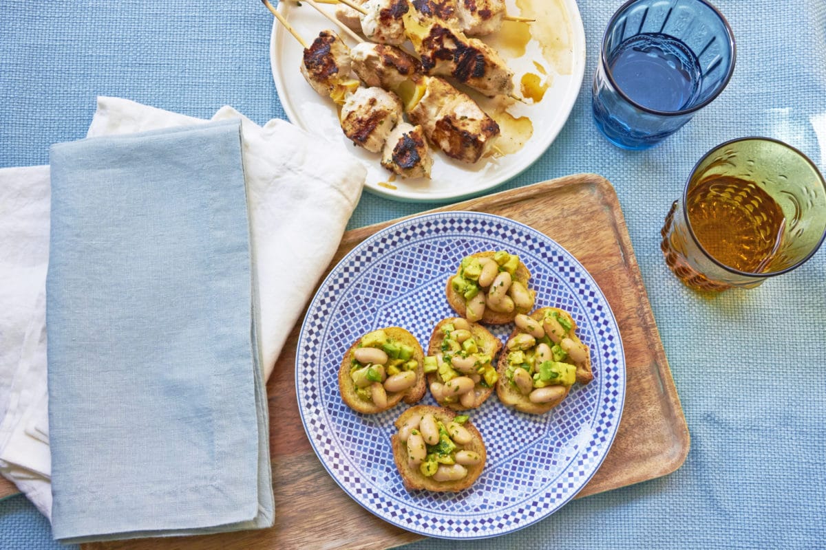 Plate of Avocado And Cannellini Bean Crostini With Gremolata next to a plate of kebabs.