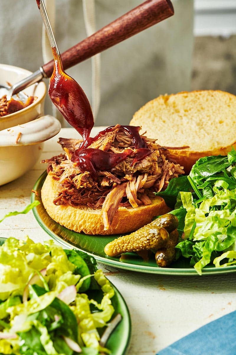 Spoon drizzling barbeque sauce onto a pulled pork sandwich.