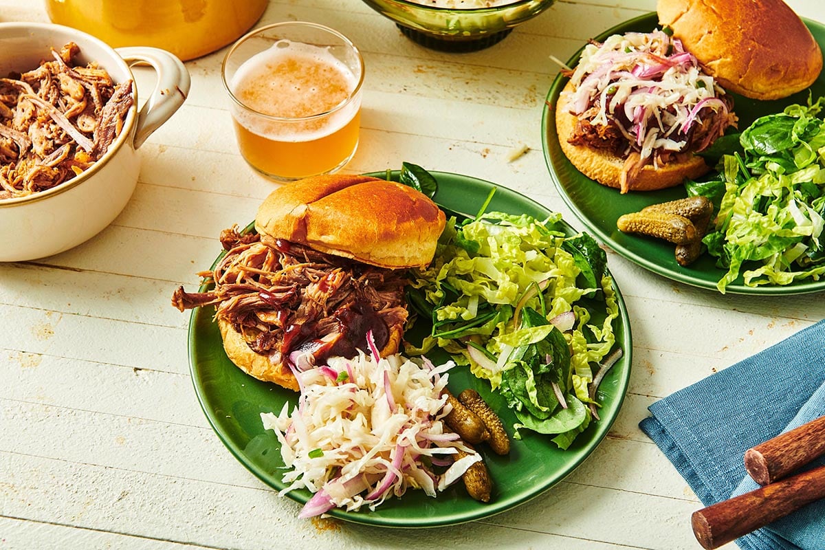 Green plate with a pulled pork sandwich, coleslaw, salad, and pickles.