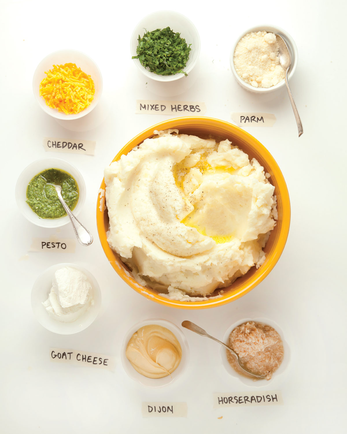 Bowl of Mashed Potatoes surrounded by containers of toppings including pesto, cheddar, and goat cheese.