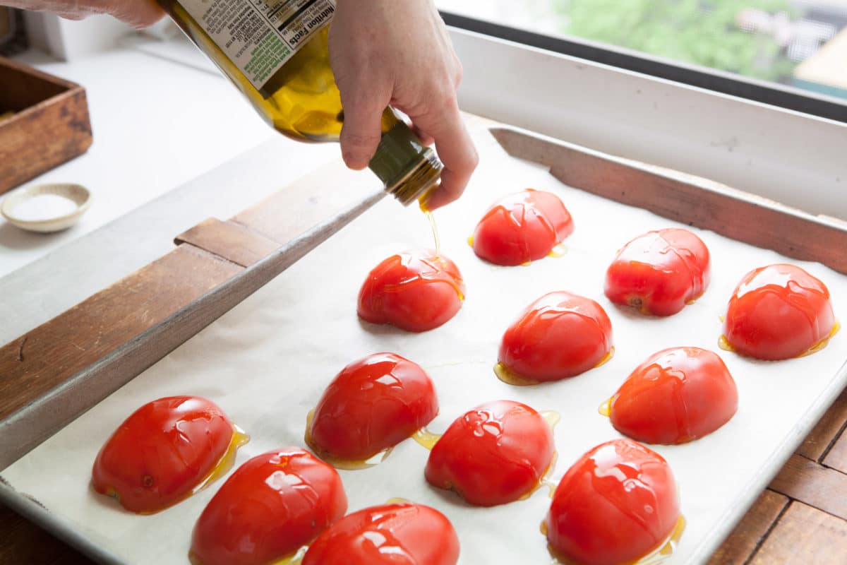 Olive oil drizzling over halved tomatoes on a lined baking sheet.