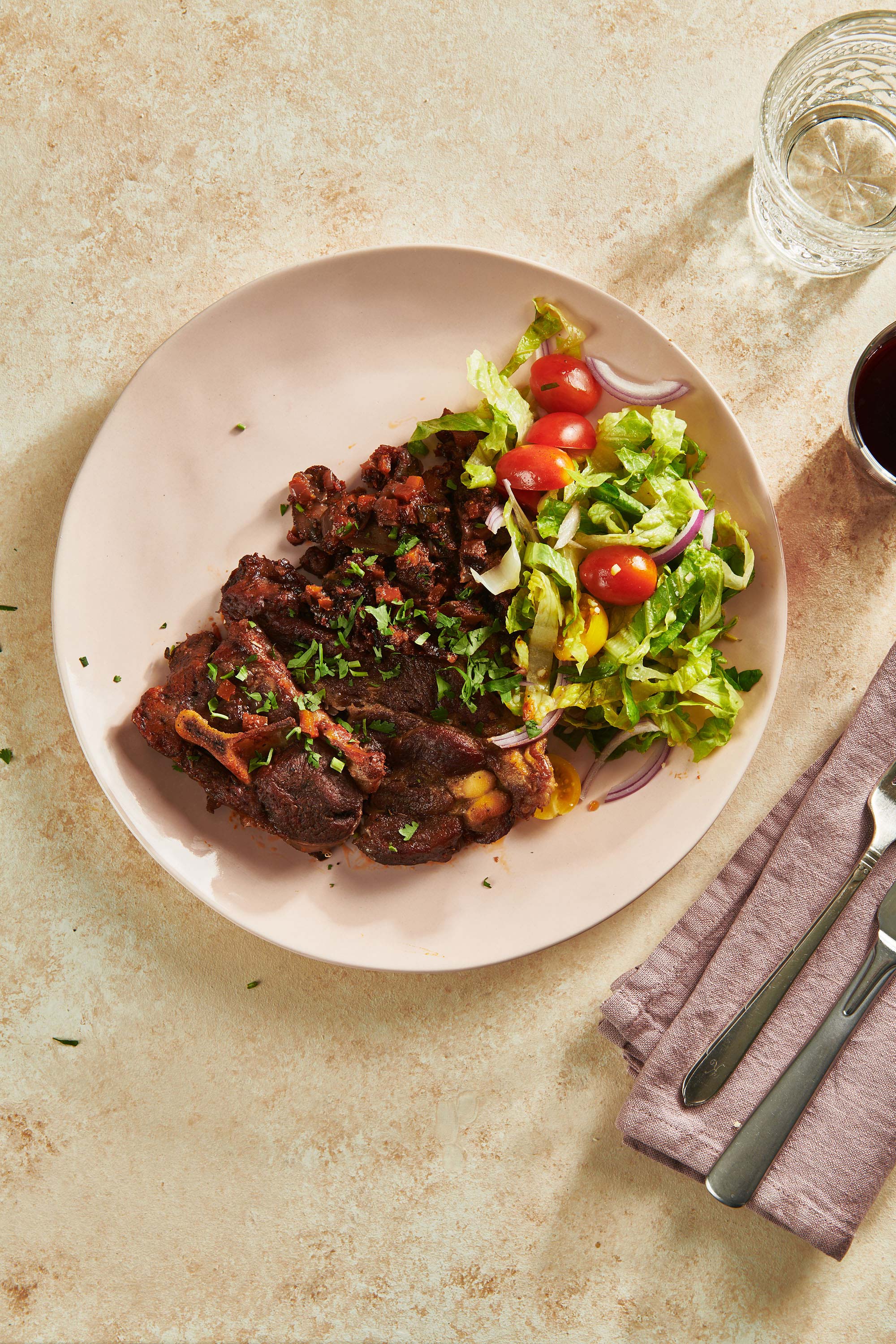 Plate with Braised Lamb Shoulder Chops and salad set with utensils and a cloth napkin.
