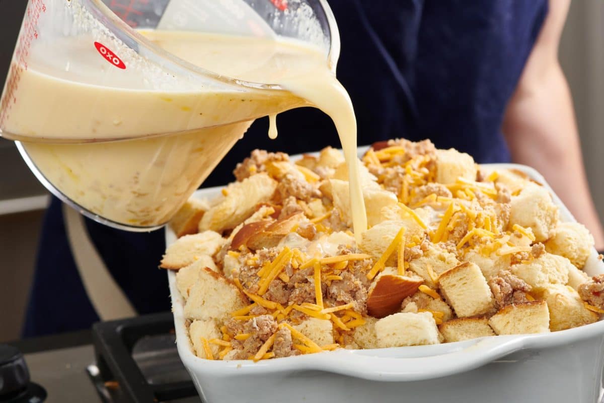 Milk mixture pouring over sausage, cheese, and Challah in a baking dish.