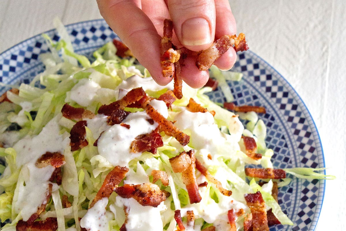 Woman sprinkling bacon onto a slivered wedge salad with buttermilk dressing.