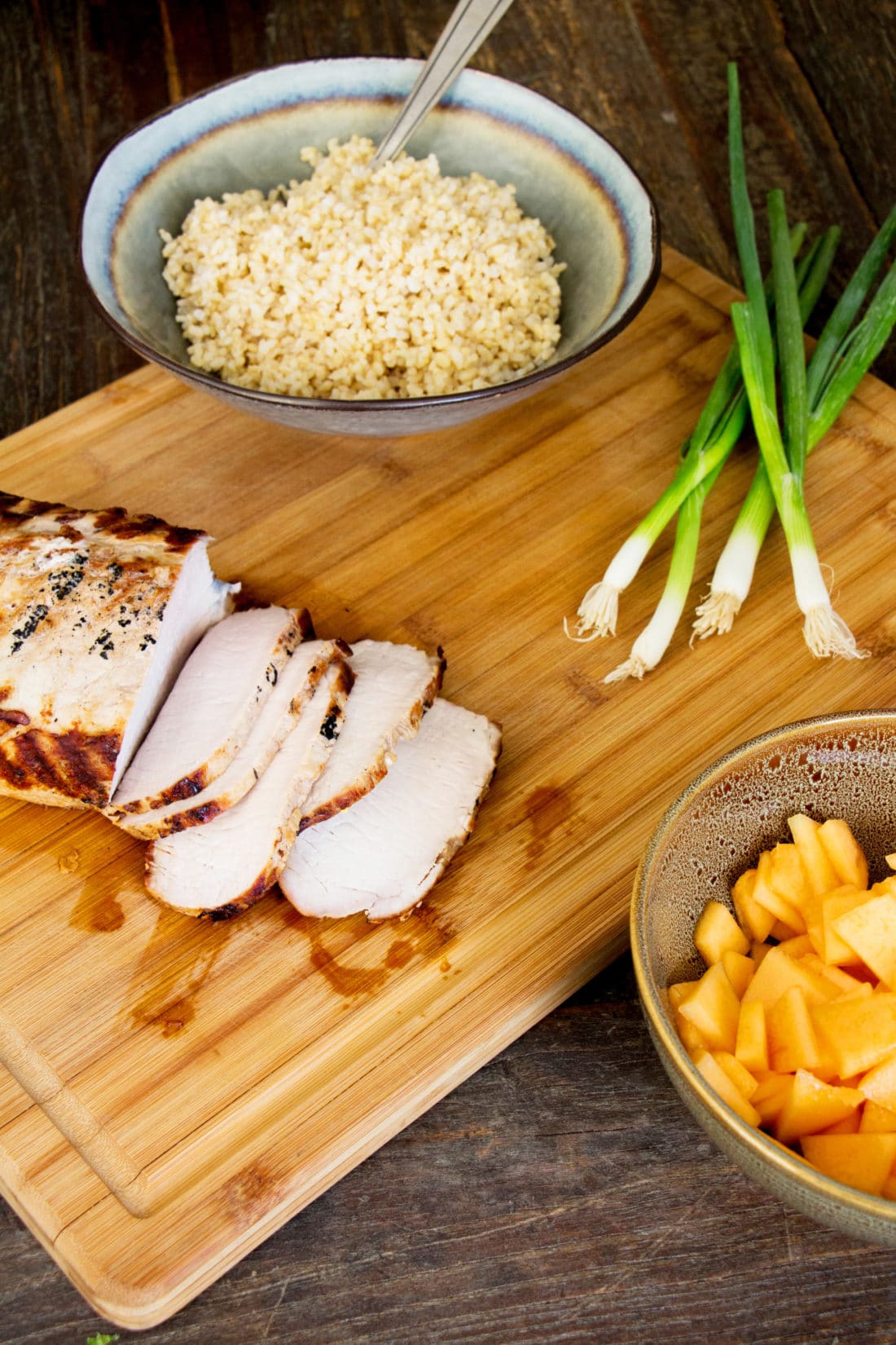 Grilled Pork Loin and ingredients for Brown Rice Salad on a wooden cutting board.