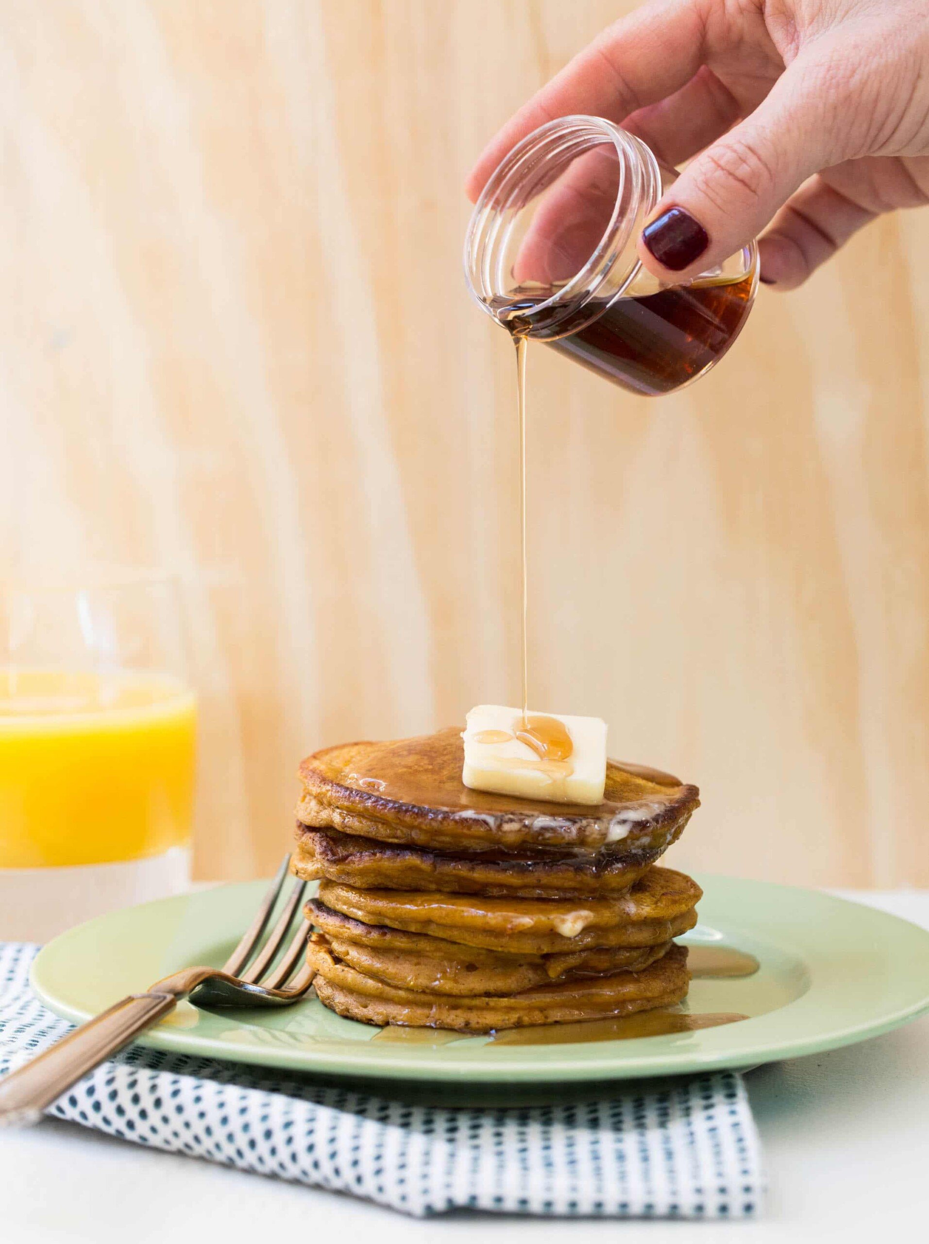 Woman pouring maple syrup over stack of pumpkin pancakes on plate.