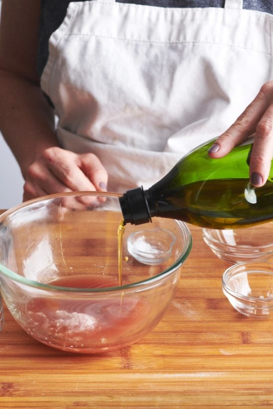 Woman pouring olive oil into a glass bowl of vinaigrette ingredients.