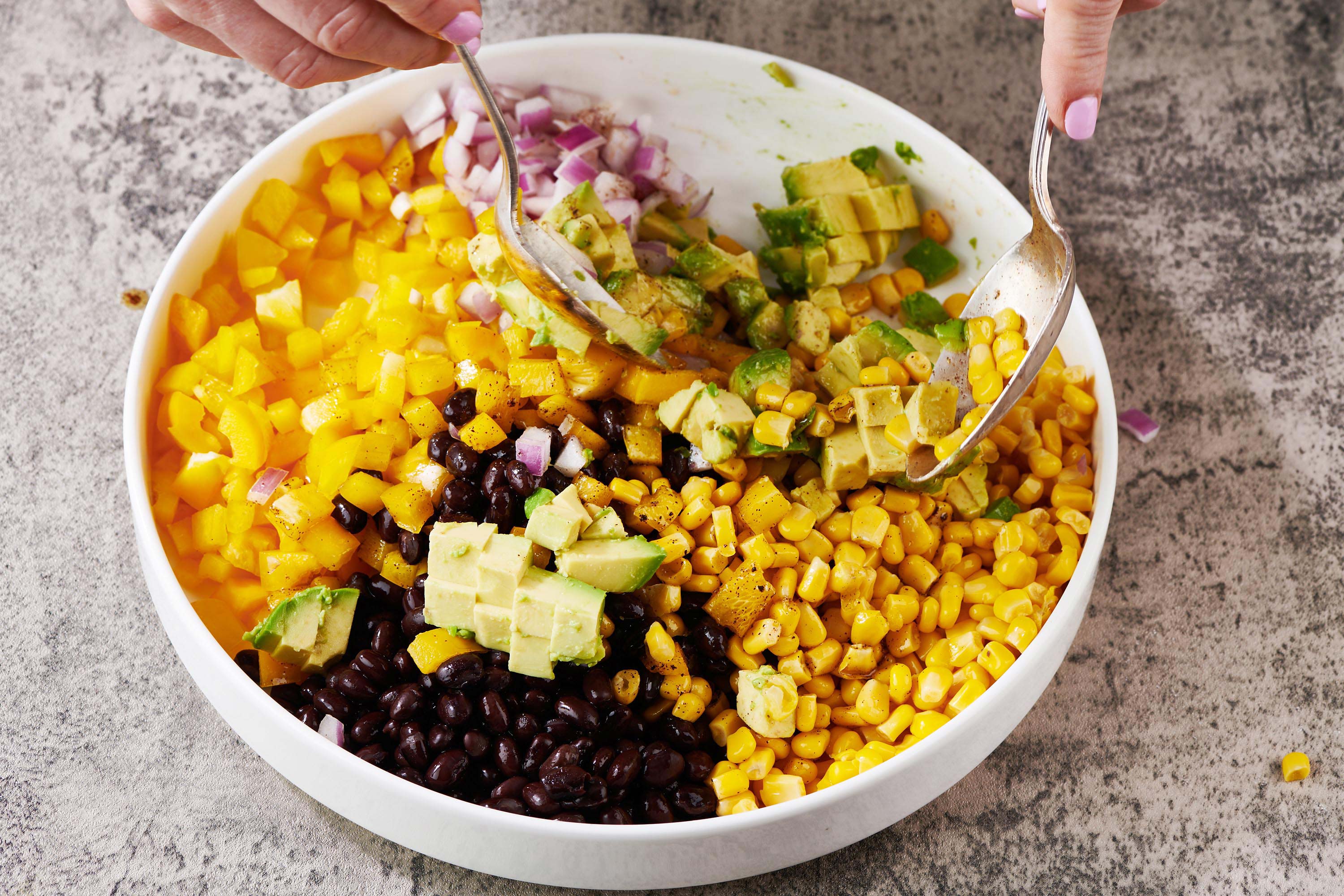 Woman using spoons to mix red onion, avocado, corn, and other ingredients for cowboy caviar.