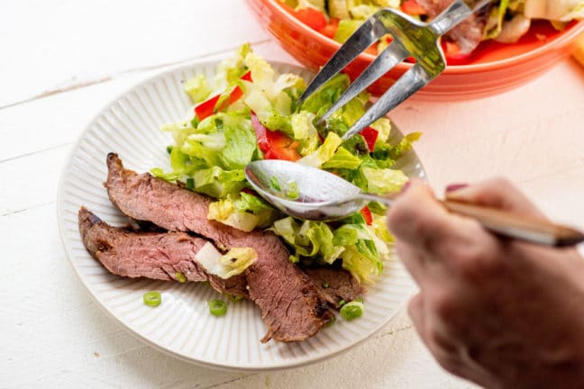 Fork and spoon serving salad onto a plate with Smoky Sweet Thai Grilled Flank Steak.