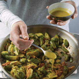 Woman adding mustard vinaigrette to a pan of brussels sprouts.