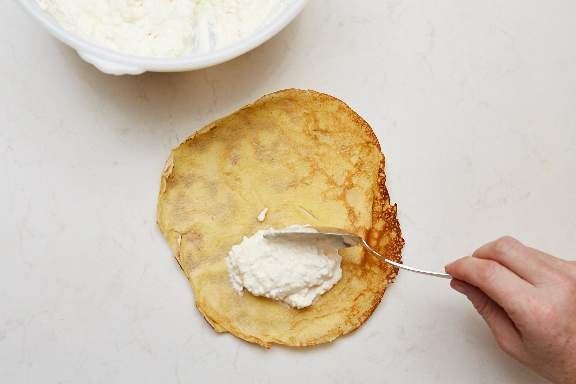 Spoon putting cheese filling on one side of a crepe.