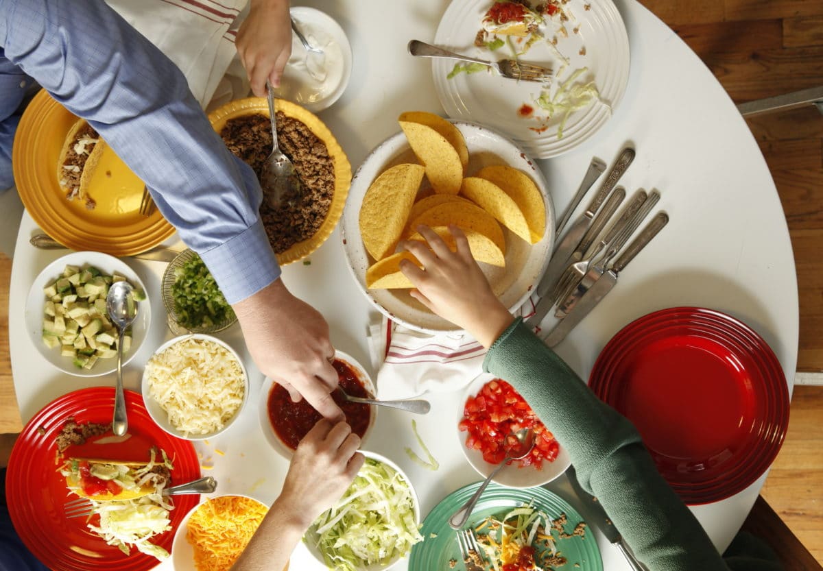 People reaching for various things at a table set with taco fillings.