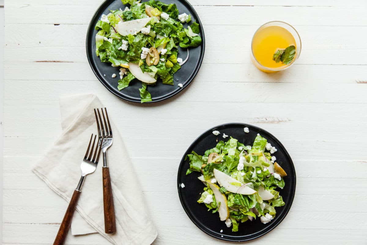 Black plates with Romaine, Pear and Goat Cheese Salad on white wood table.