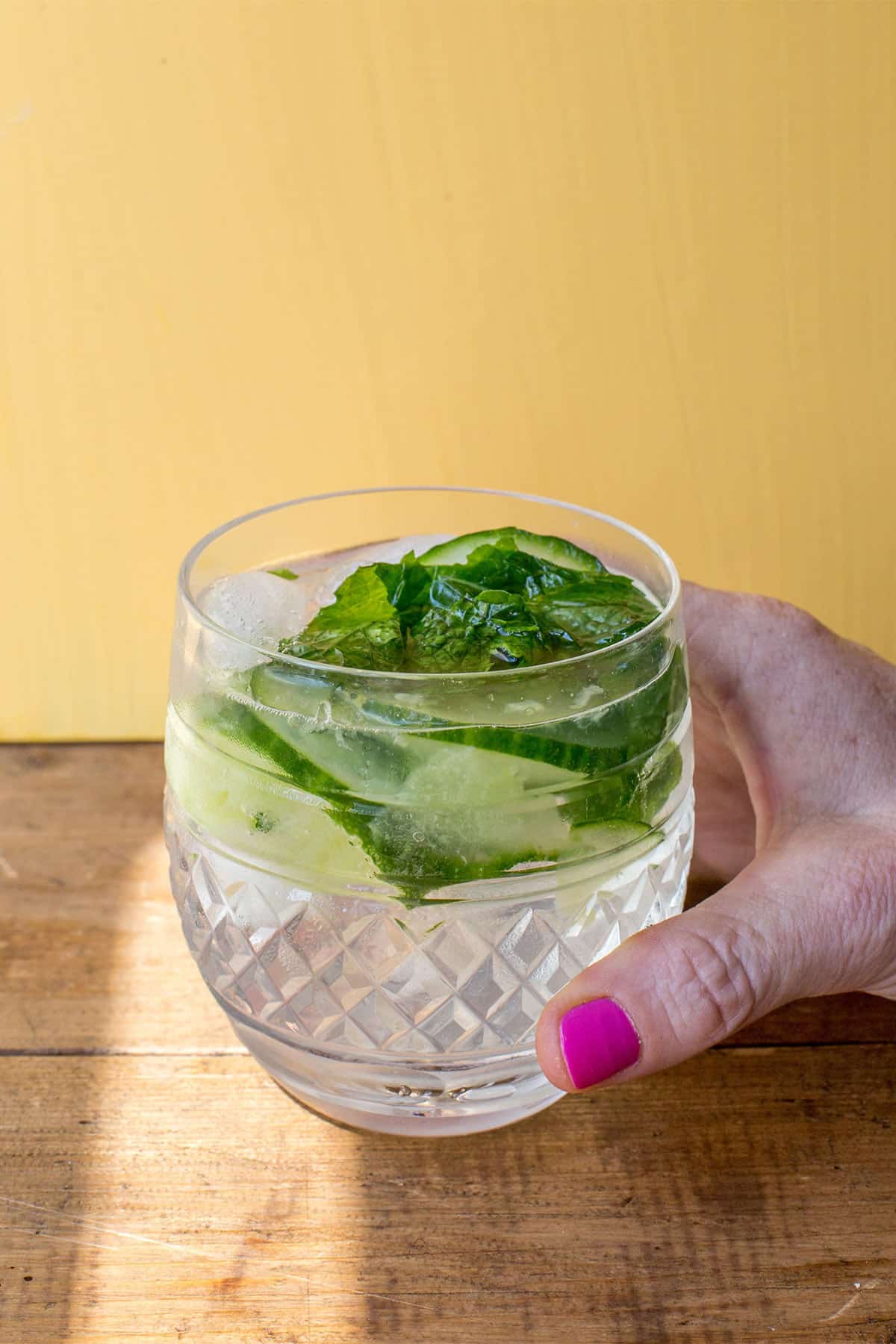 Woman holding a Gin and Tonic with mint and cucumber over wood board.