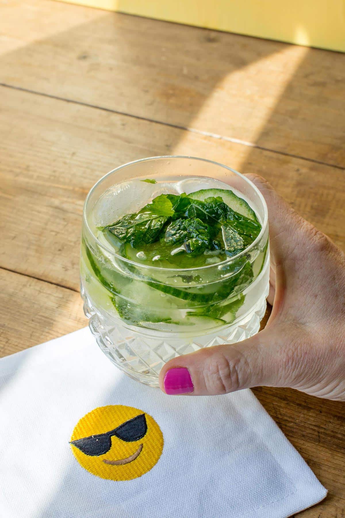 Woman holding a fresh Cucumber and Mint Gin and Tonic glass over a smiley face napkin.