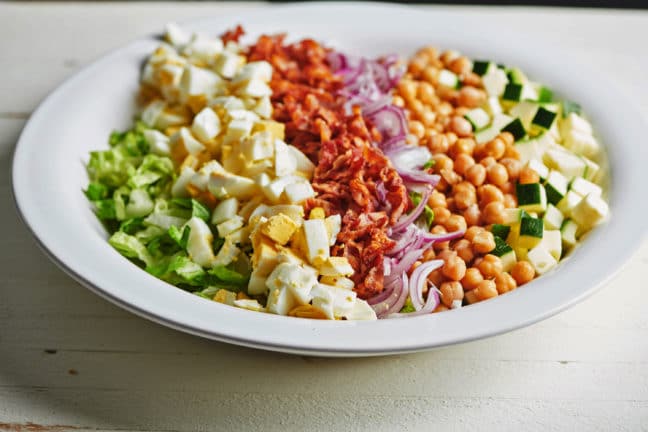 Large plate of hard boiled egg, bacon, red onion, and chickpeas over lettuce and zucchini.