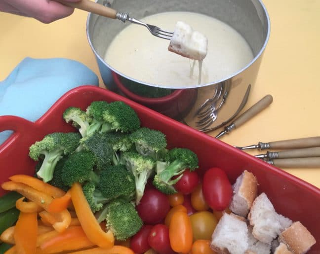 Fork dipping bread into Classic Swiss Fondue next to a tray of veggies.