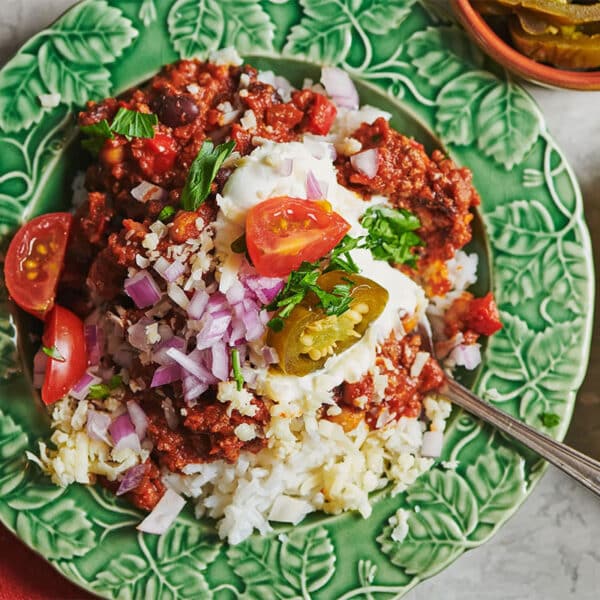 Chili over rice in green bowl topped with fresh onions, tomatoes, and jalapeños.