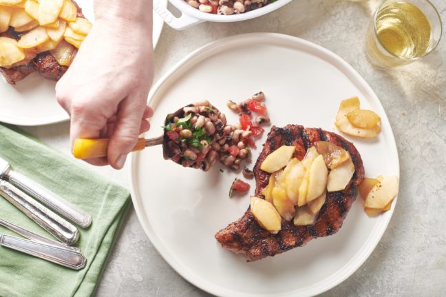 Person spooning black eyed pea salad onto a plate with Pork Chops with Apples.