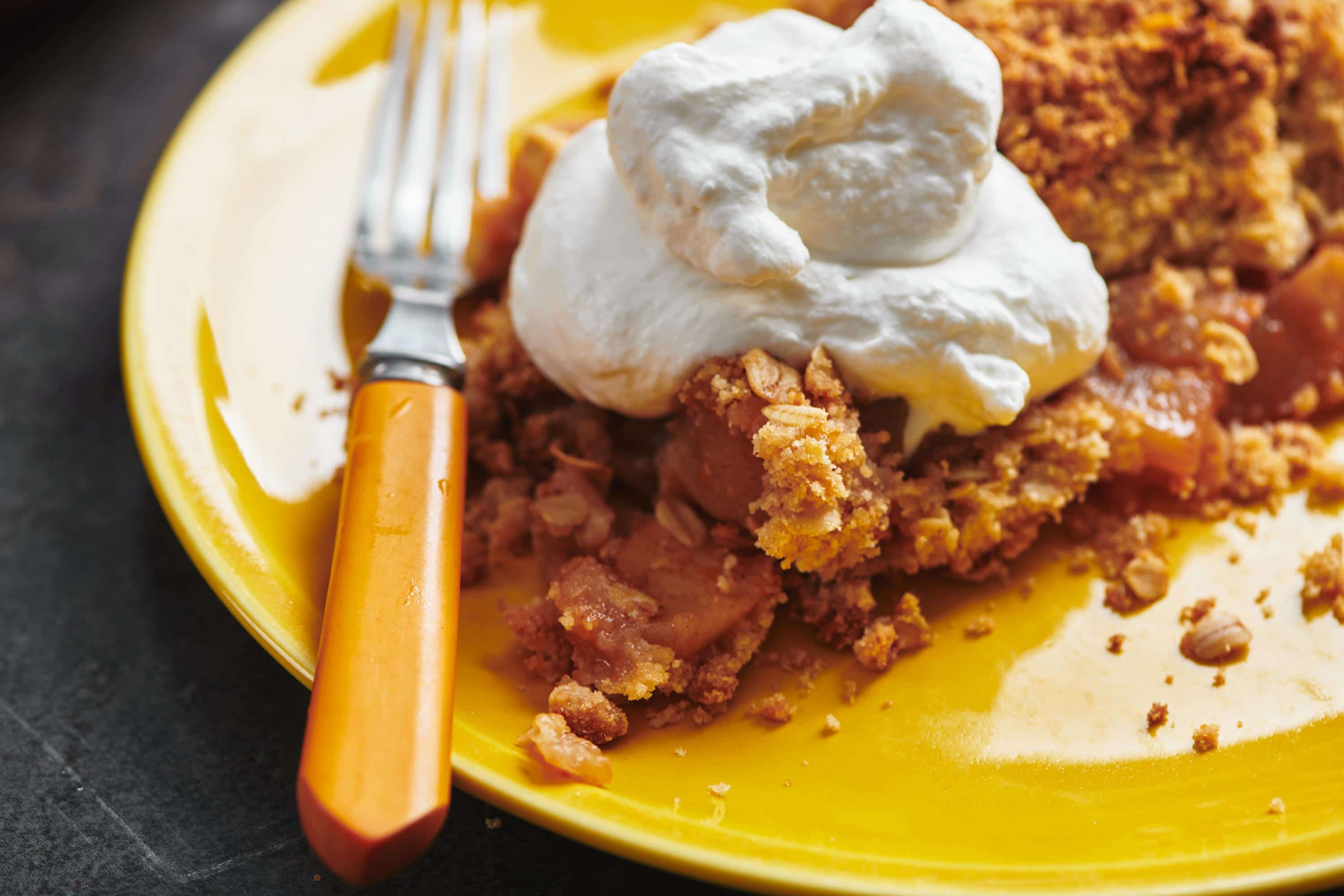 Apple crisp topped with whipped cream on yellow plate with fork.