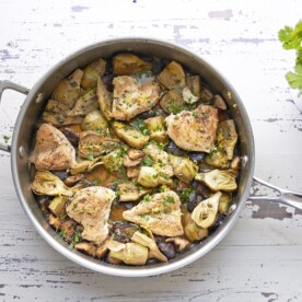 Skillet of Braised Chicken, Mushrooms and Baby Artichokes set on a white, wooden table.