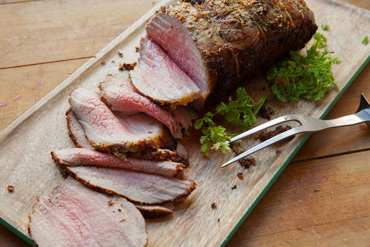 Sliced eye of round roast with thyme and rosemary on cutting board.