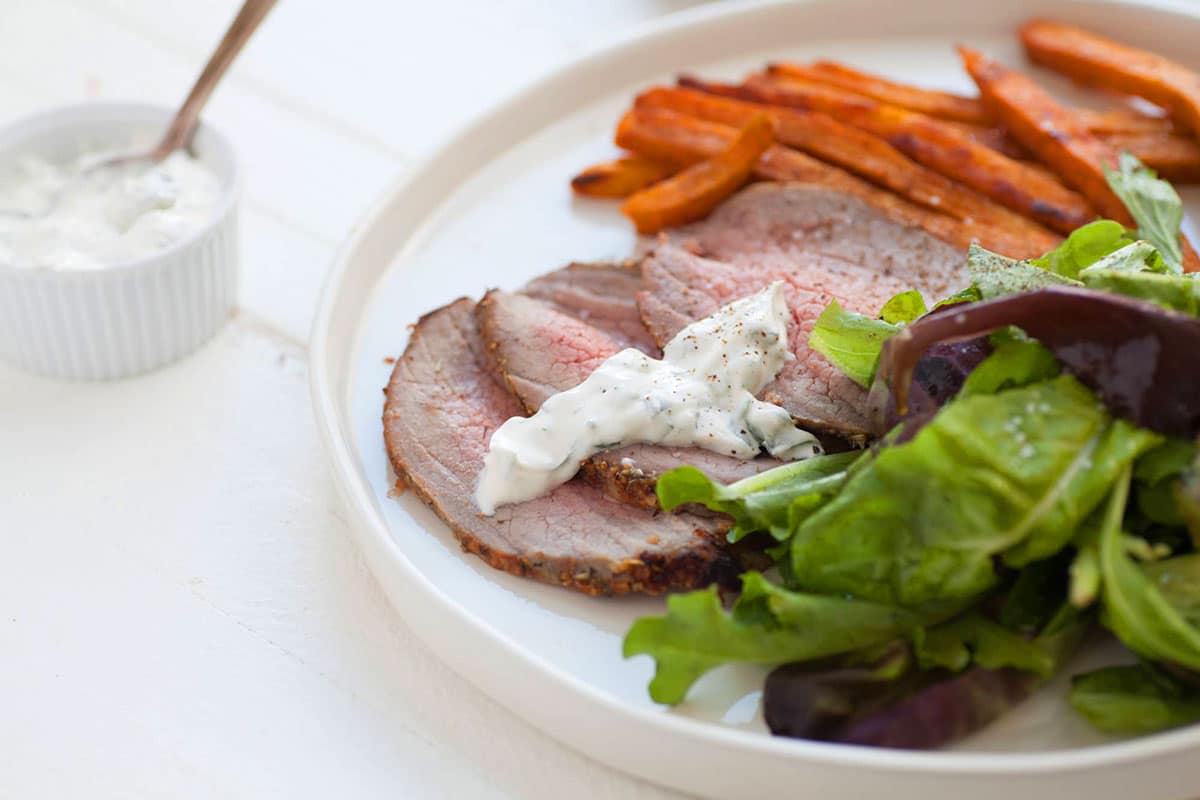 Sliced eye of round with herb sauce on plate with salad and carrots.