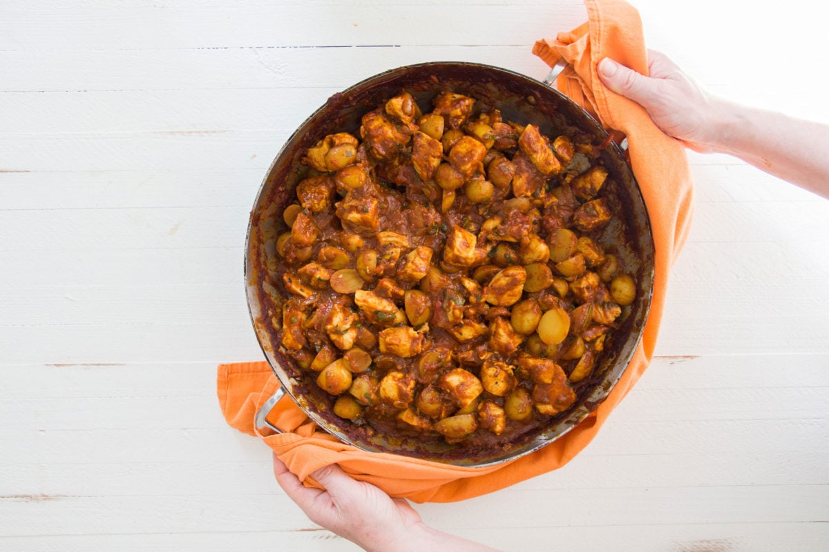 Woman using an orange cloth to grab a pan of Indian Spiced Chicken and Potato Stew.