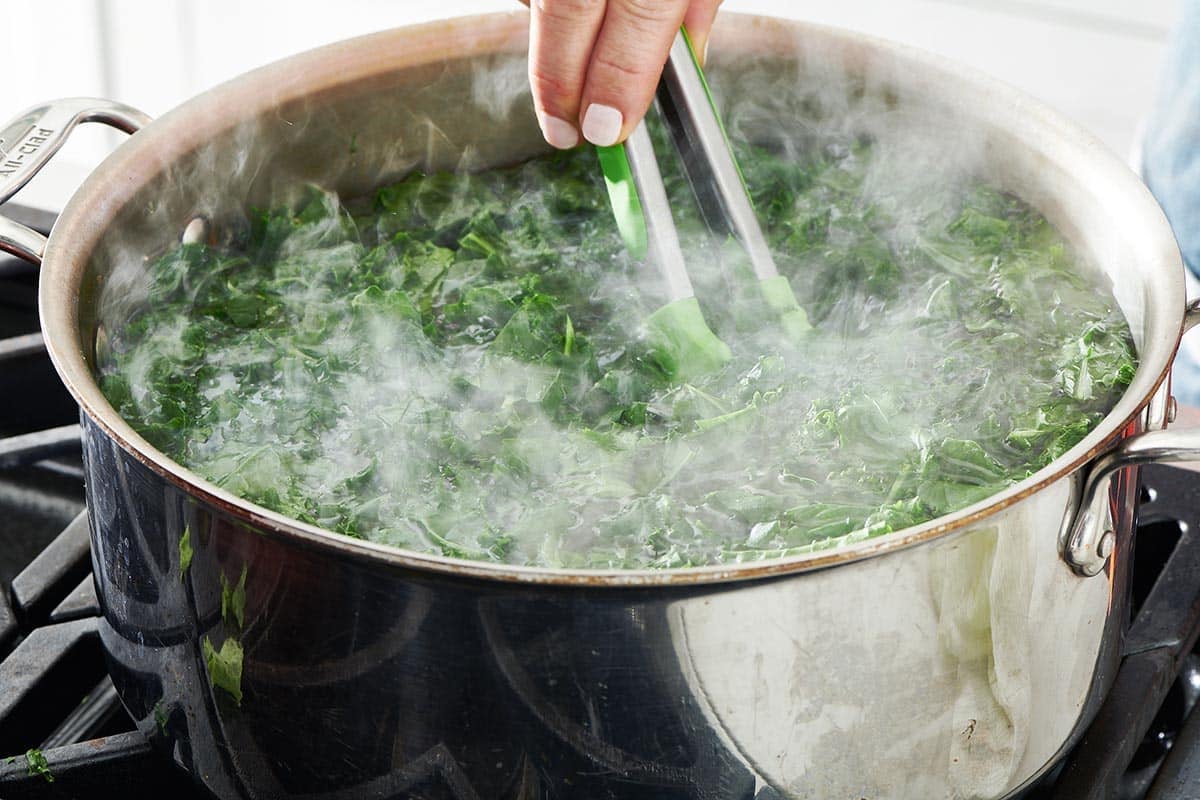 Woman using tongs in a steaming bowl of kale.