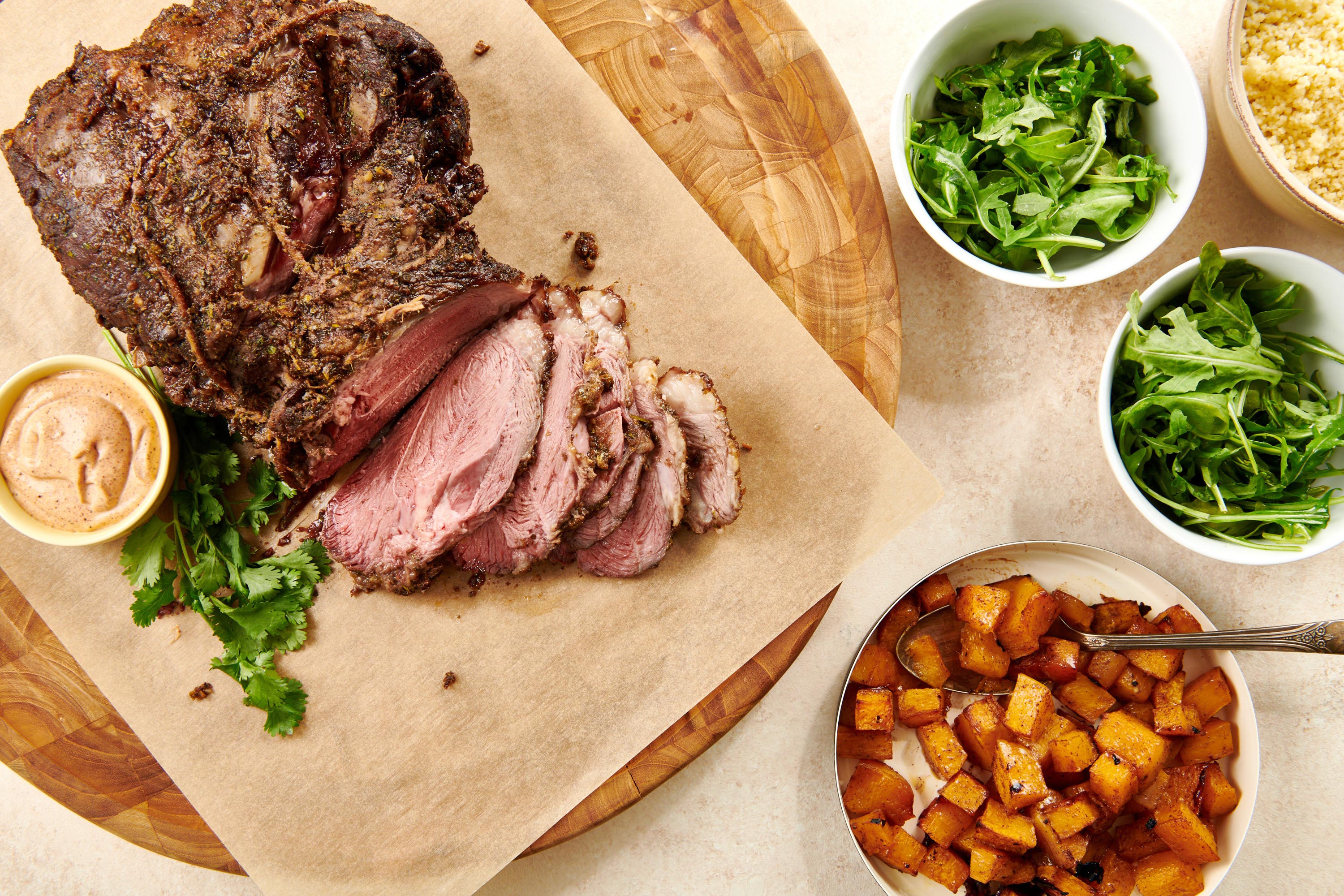 Moroccan Leg of Lamb on a parchment-lined wooden board with bowls of sides.