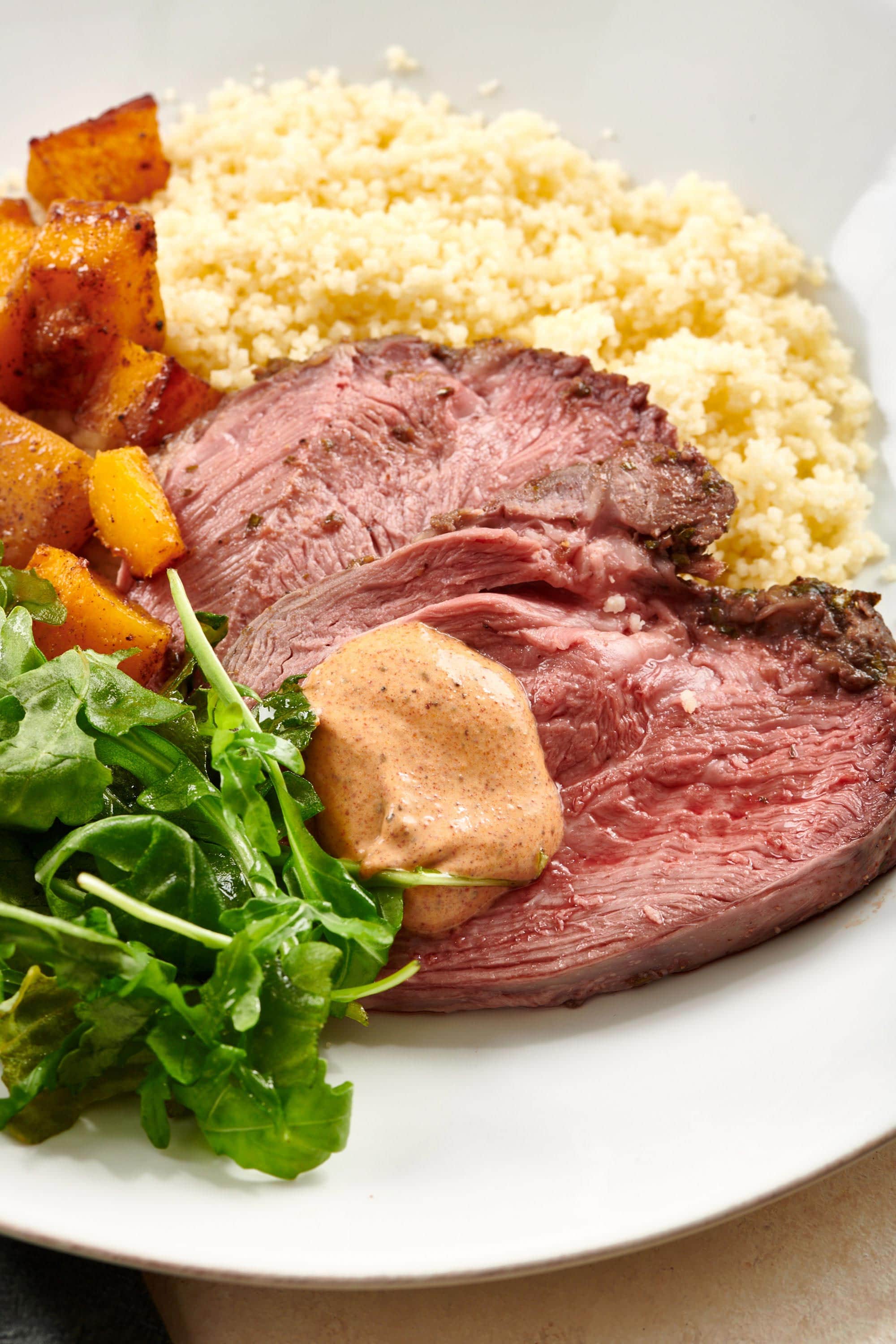 Sliced Lamb topped with harissa sauce and on a plate with arugula, squash, and couscous.