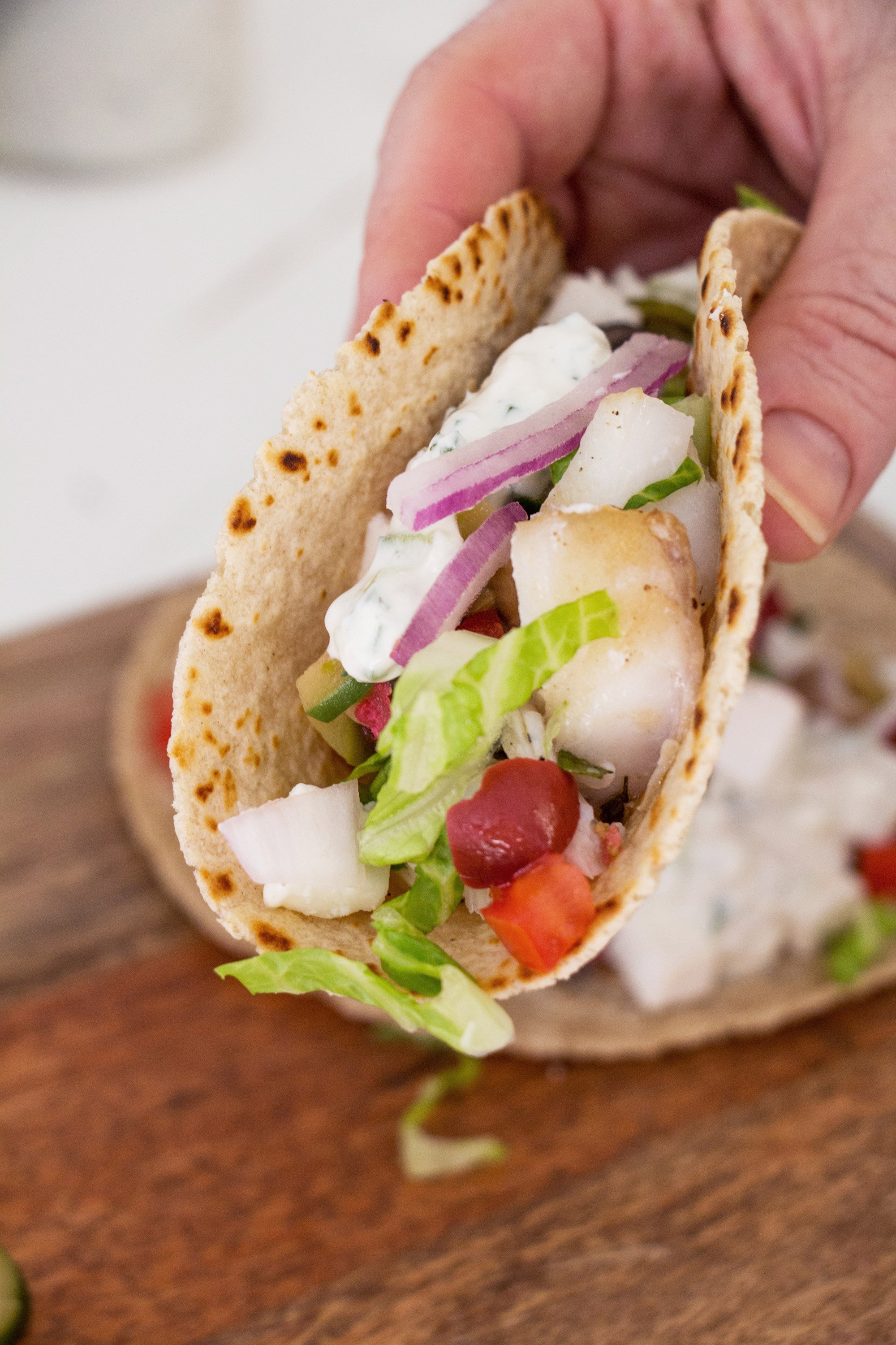 Person holding a Greek Fish Taco stuffed with ingredients such as feta and red onions.