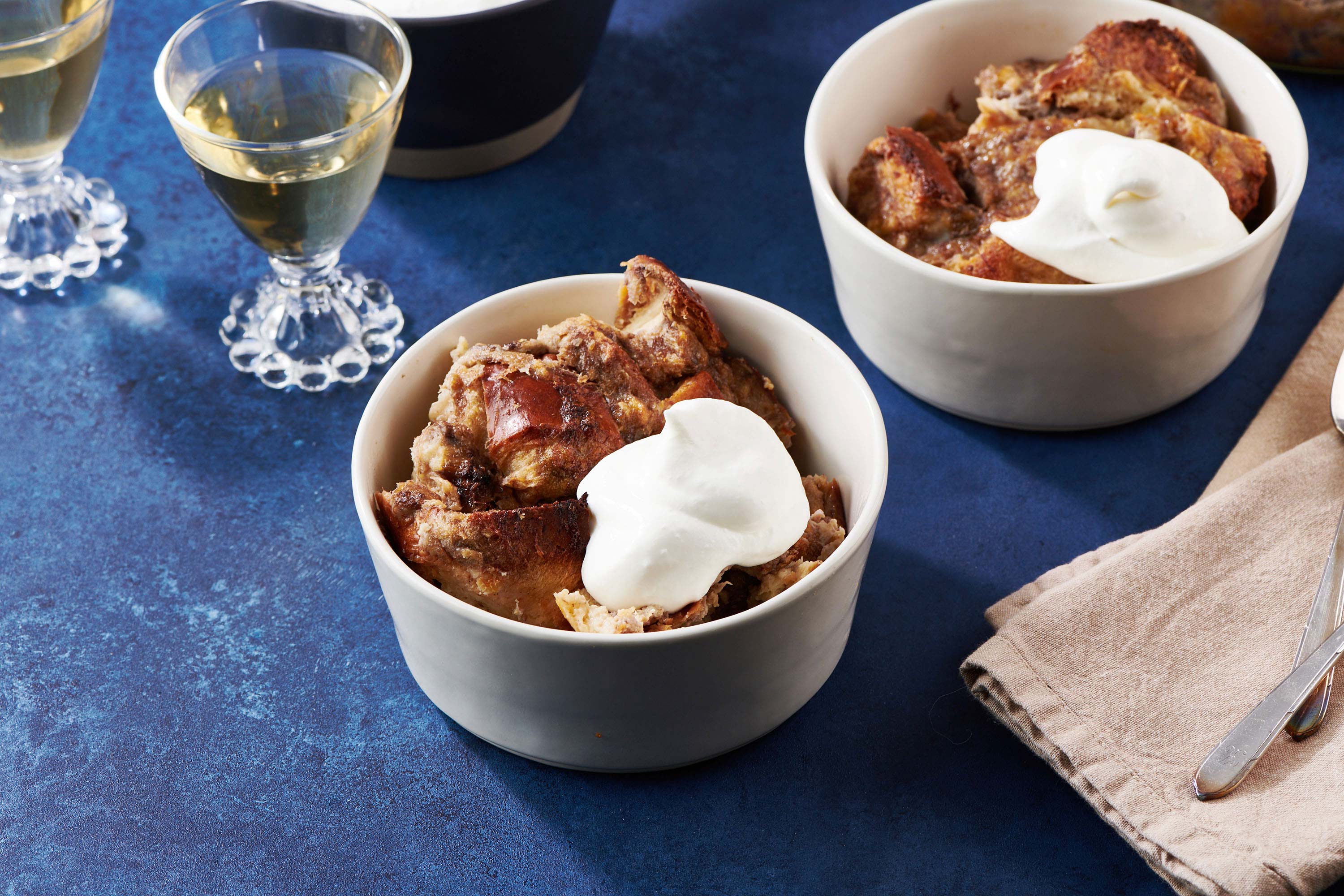 Two bowls of Banana Bread Pudding topped with whipped cream with small glasses of white wine.