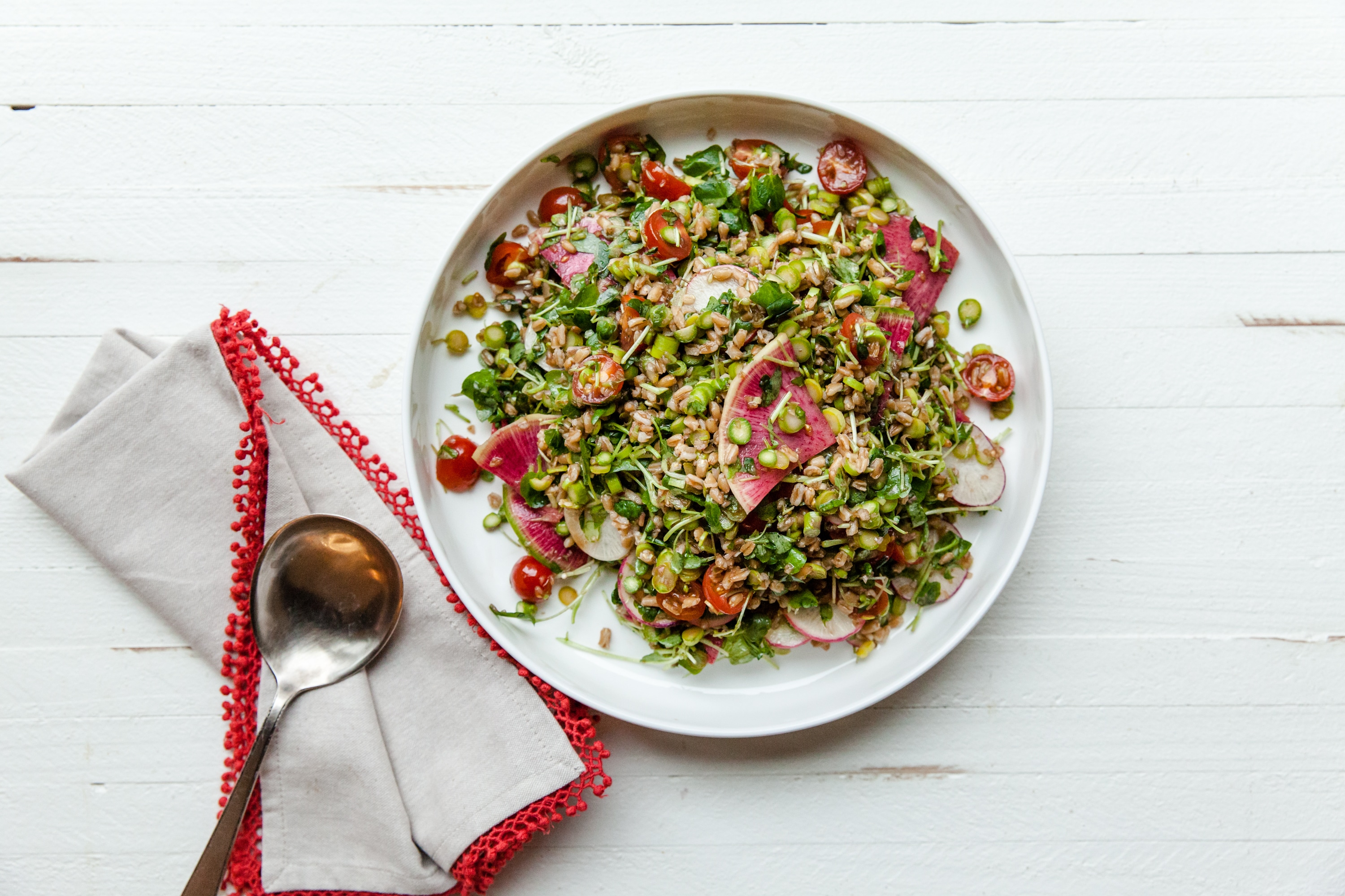 Farro and Vegetable Salad in a white serving bowl on a white table with a spoon on a napkin with red trim.
