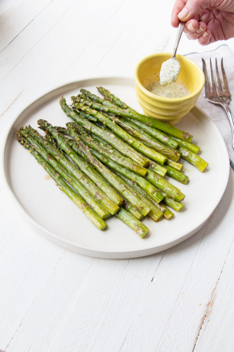 Bowl of Creamy Mustard-Oregano Sauce on a plate of Roasted Asparagus.