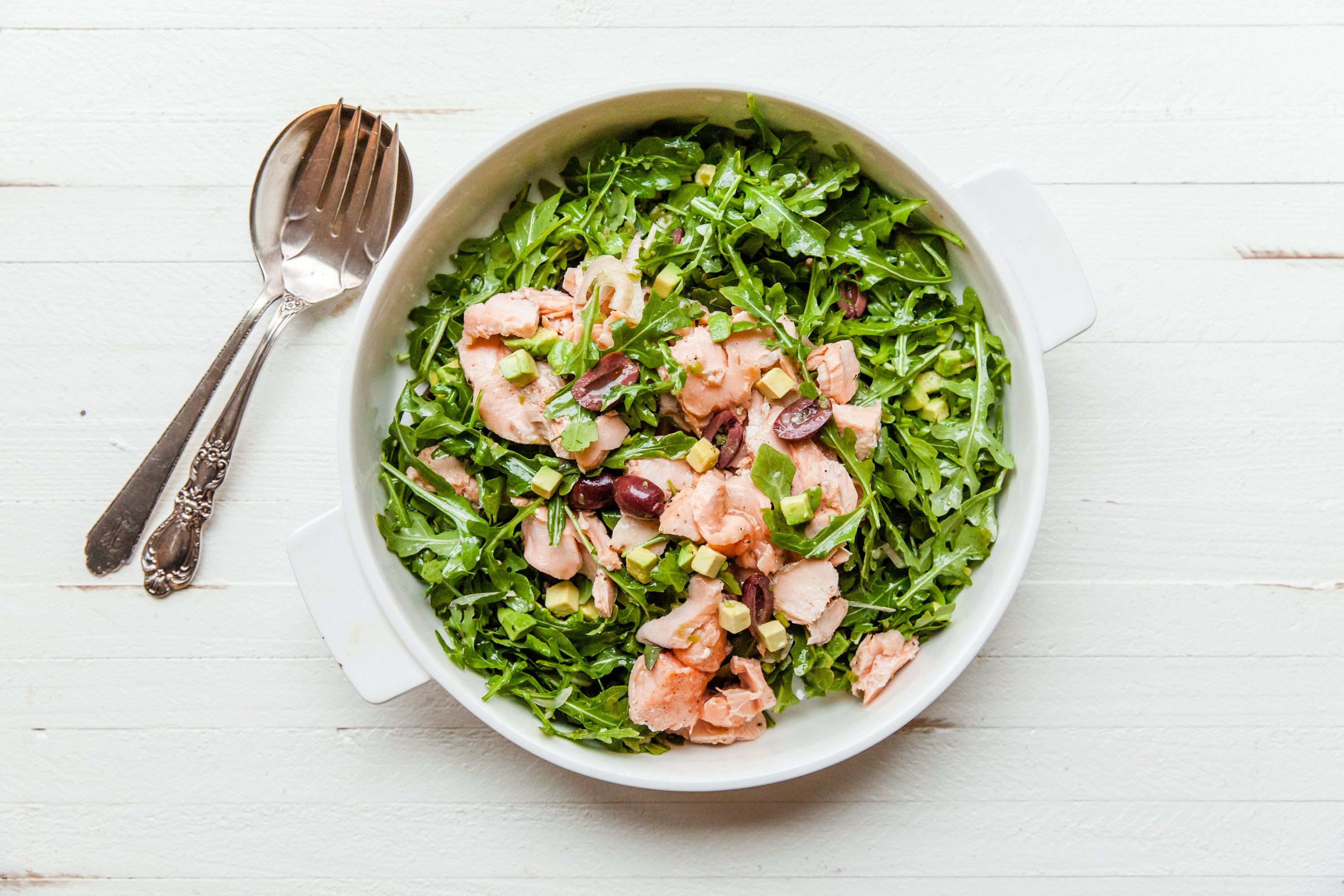 Salmon, Arugula, and Avocado Salad  on a white table with serving utensils.