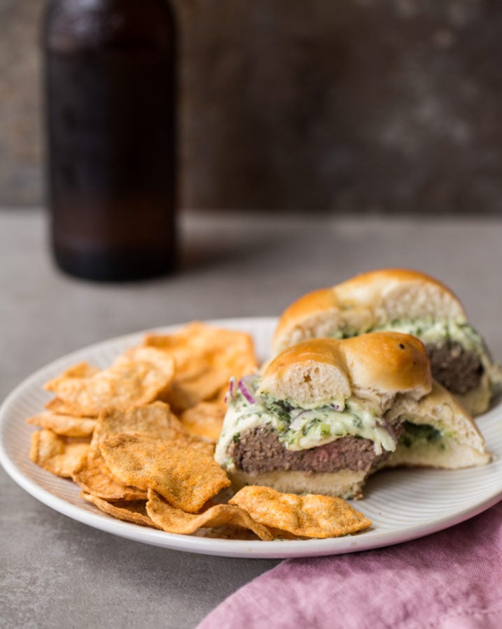 Halved Grilled Cheeseburger with Herb Sauce missing a bite on a plate with chips.