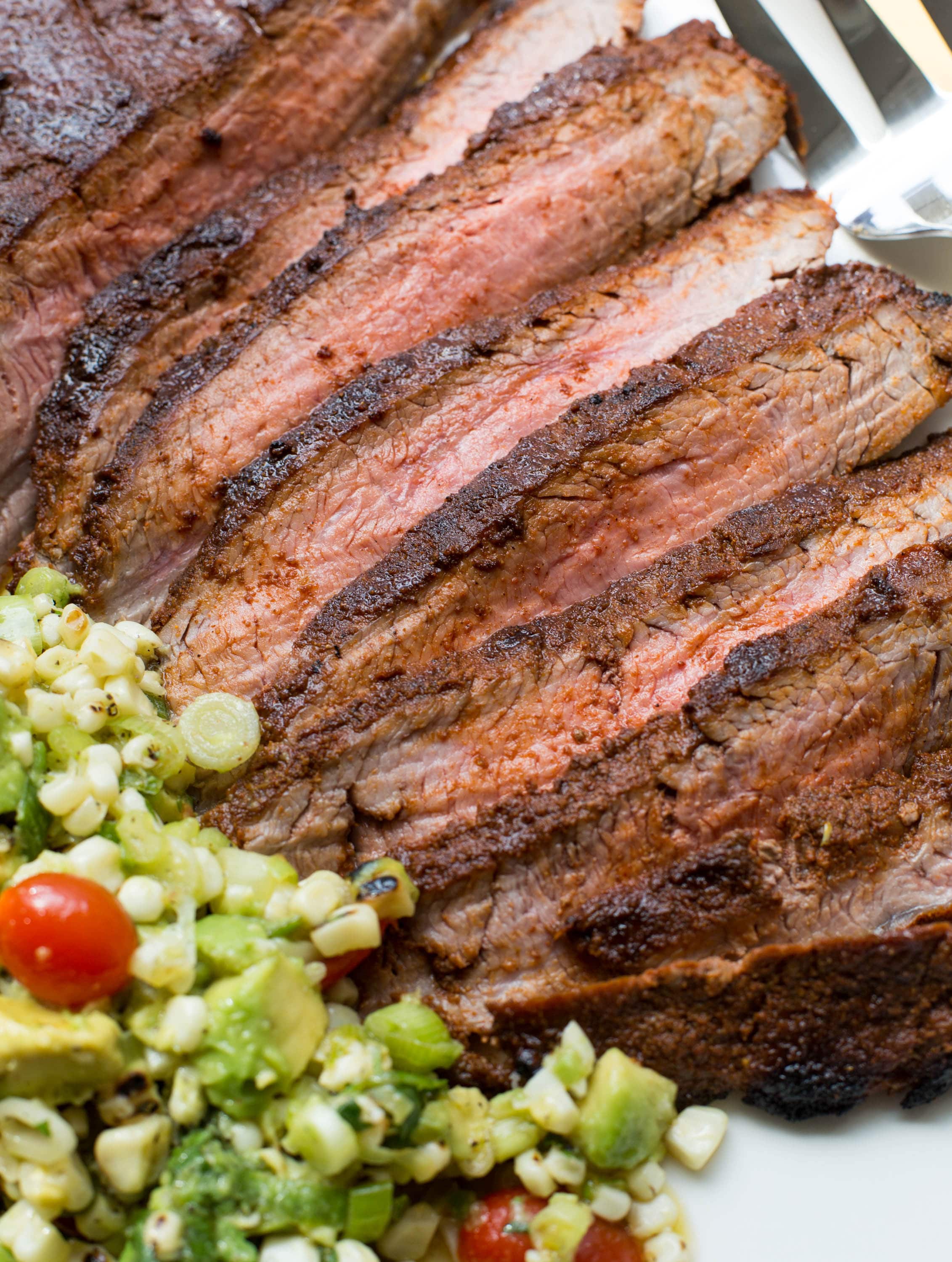 Slices of Chili Rubbed Flank Steak on a tray with Corn, Tomato and Avocado Salad.