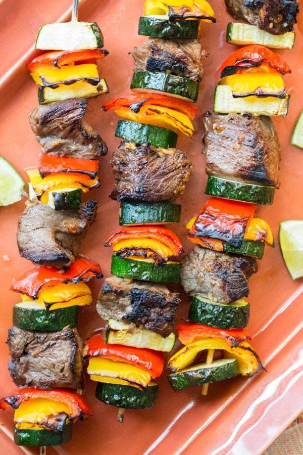 Three Beef Teriyaki Kebabs with Peppers and Zucchini on a tray with cut limes.