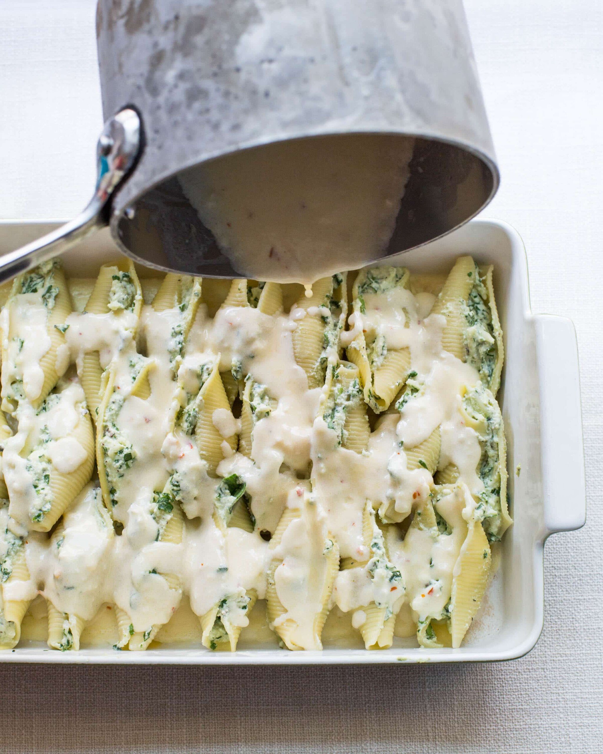 Gouda cheese sauce pouring over a baking dish of Spinach and Cheese Stuffed Shells.