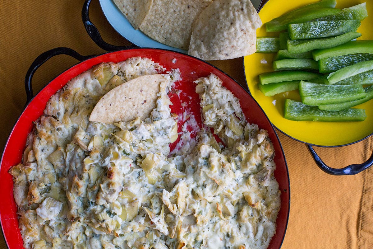 Spicy Cheesy Artichoke Dip in red bowl with chips and peppers.