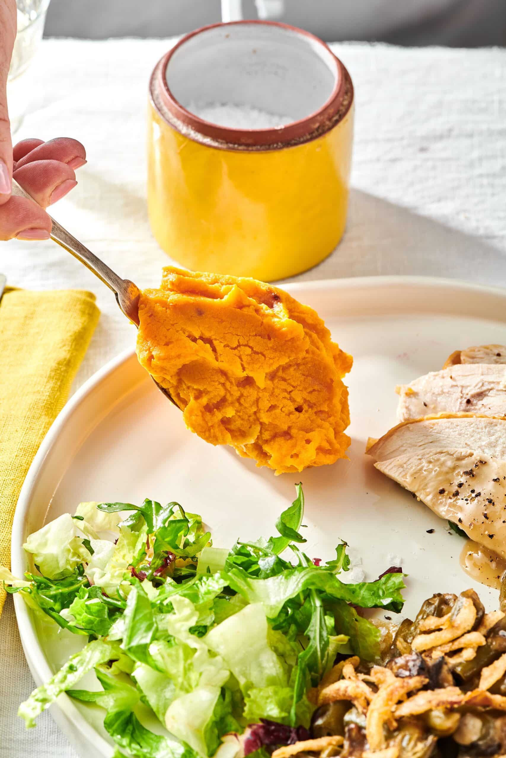 Spoon scooping Garlic Mashed Sweet Potatoes onto a plate with meat and salad.