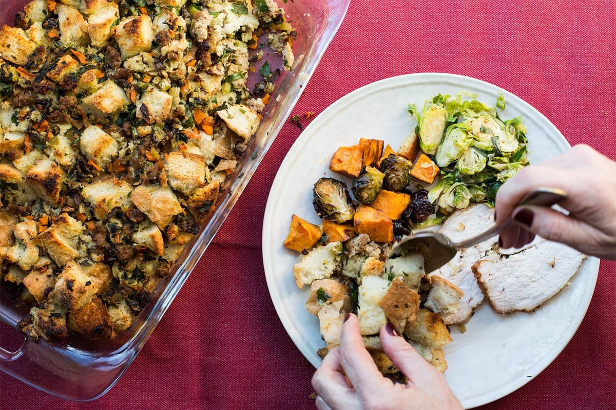 Woman adding Bread Stuffing with Turkey Sausage to a plate with casserole of stuffing nearby.