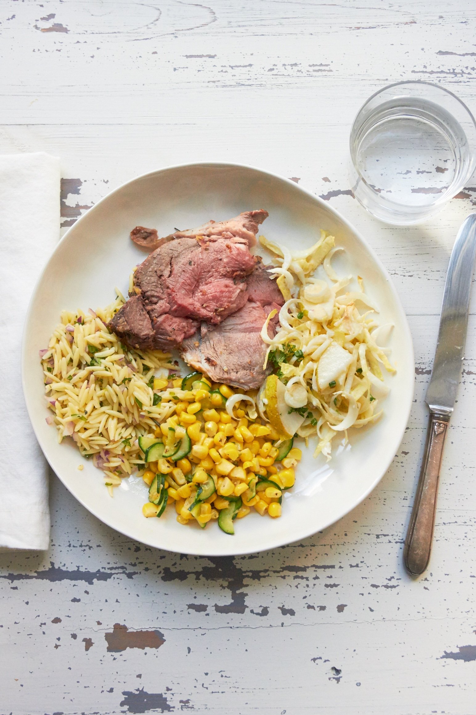 Slices of Roasted Leg of Lamb with Greek Orzo, Creamy Endive Salad and Sauteed Corn sides on plate.