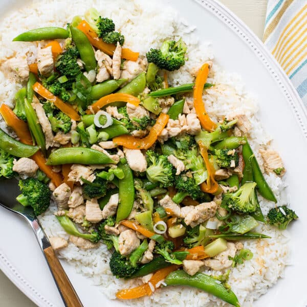 Stir-fry with chicken, broccoli, and sugar snap peas served over white rice on plate.