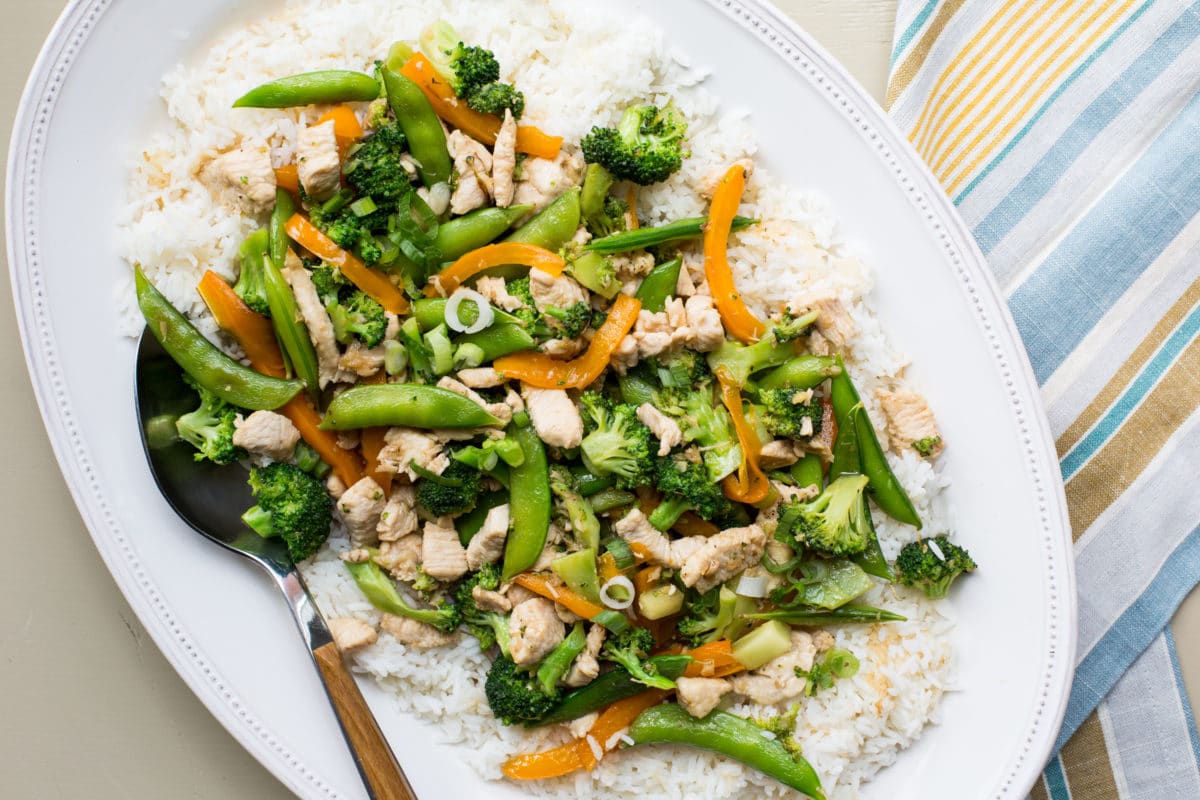 Stir-fry with chicken, broccoli, and sugar snap peas served over white rice on plate.