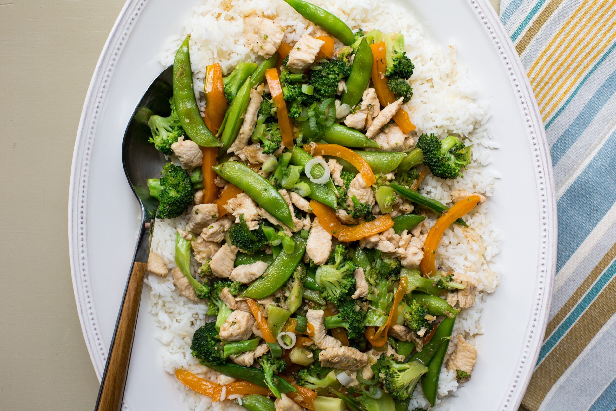 White plate with chicken, broccoli, and snap pea stir-fry over rice.
