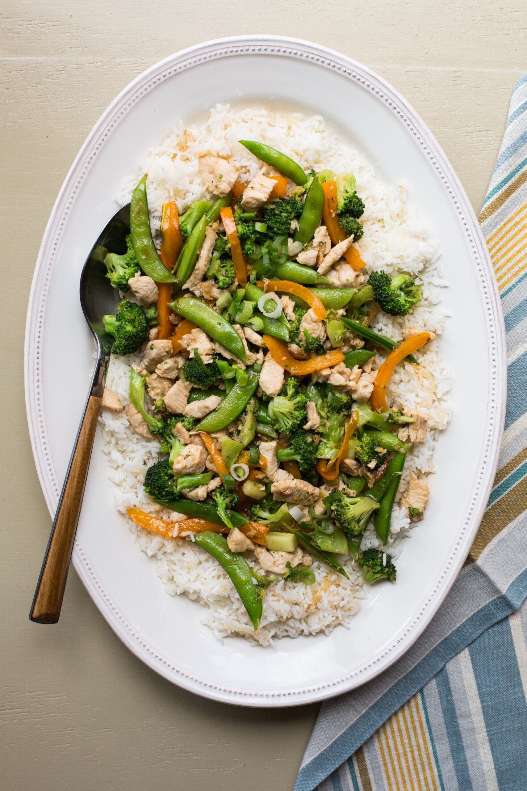 Chicken stir fry with broccoli and sugar snap peas over rice on plate.