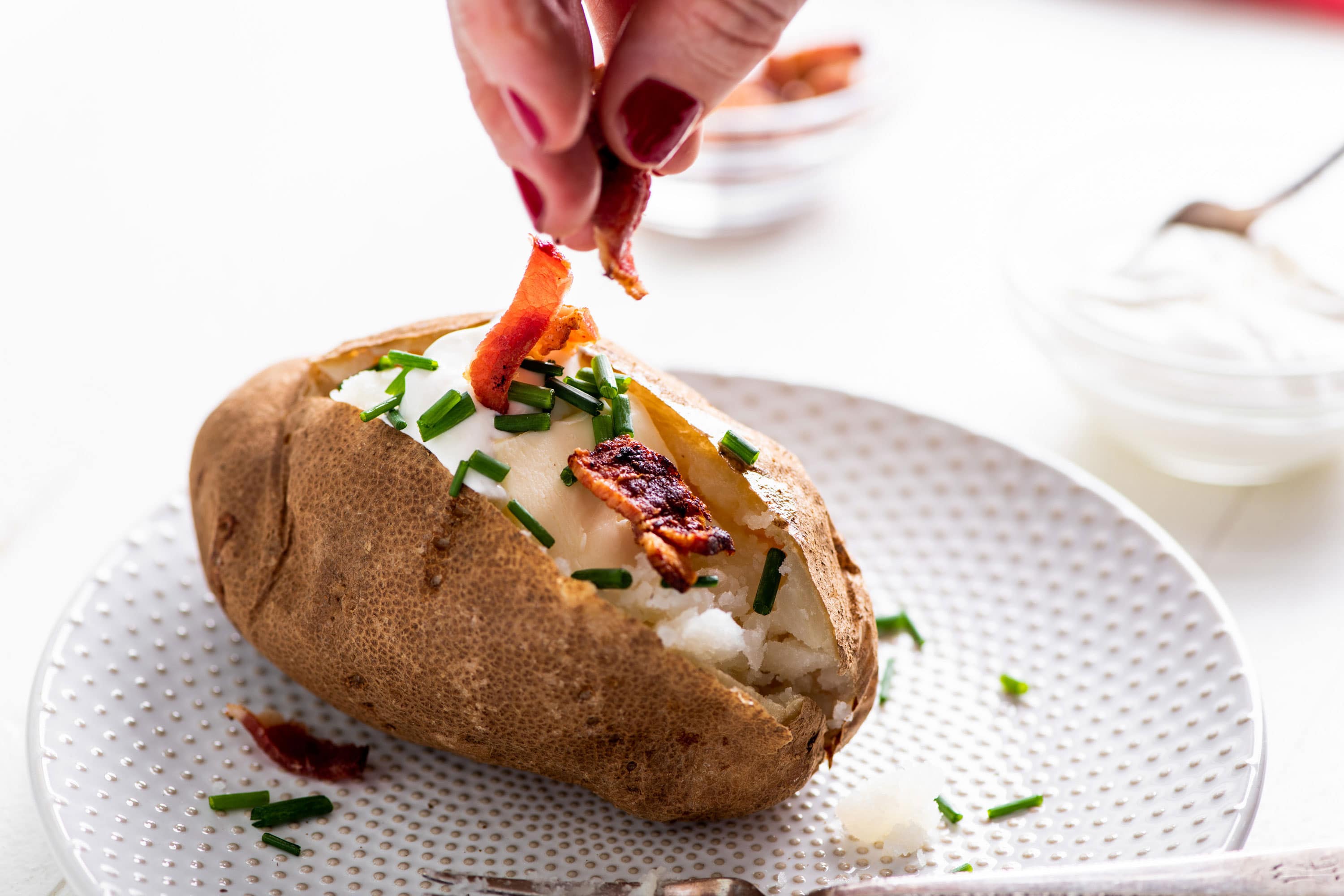 Adding bacon to baked potato with chives and sour cream on plate.