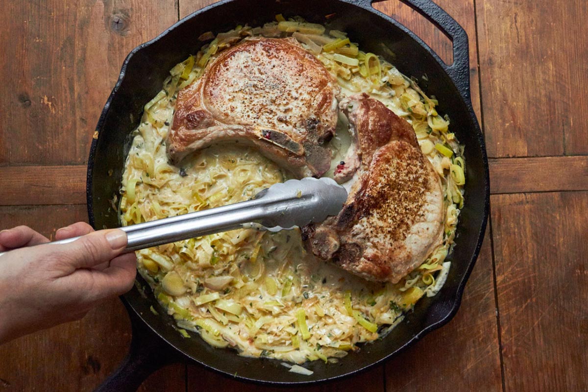 Woman using tongs to flip pork chops in creamy madeira sauce in pan.